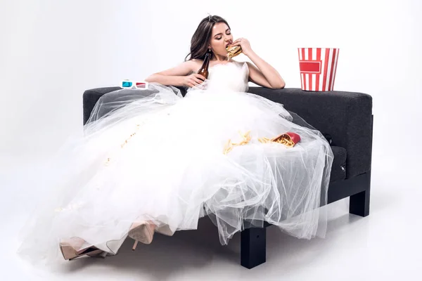 Depressive hungrige junge Braut im Brautkleid sitzt auf der Couch und isst Burger mit Bier auf Weiß — Stockfoto