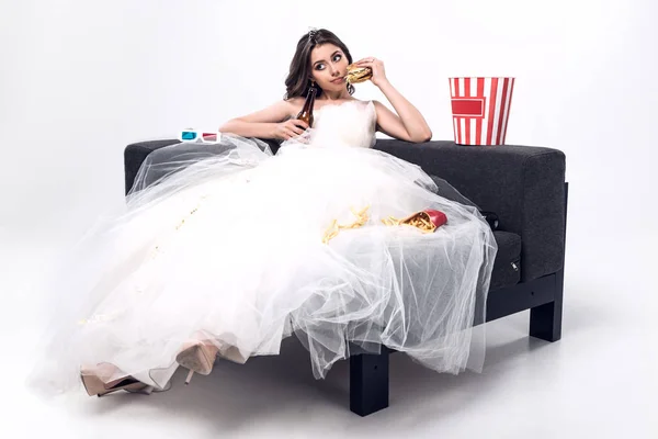 Triste jeune mariée en robe de mariée assis sur le canapé avec de la bière et de la malbouffe sur blanc — Photo de stock