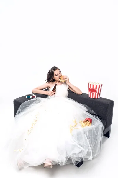 Hungry young bride in wedding dress sitting on couch with beer and junk food on white — Stock Photo
