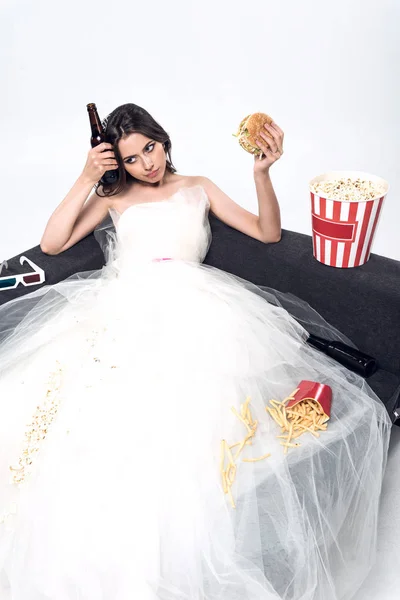 Jeune mariée déprimée en robe de mariée assise sur le canapé avec de la bière et de la malbouffe sur blanc — Photo de stock