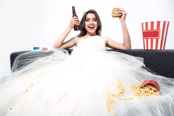 Glückliche junge Braut im Brautkleid sitzt auf der Couch mit Bier und Junk Food isoliert auf weiß — Stockfoto