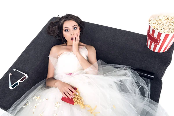 Високий кут зору шокованої молодої нареченої у весільній сукні, що сидить на дивані з нездоровою їжею ізольовано на білому — стокове фото