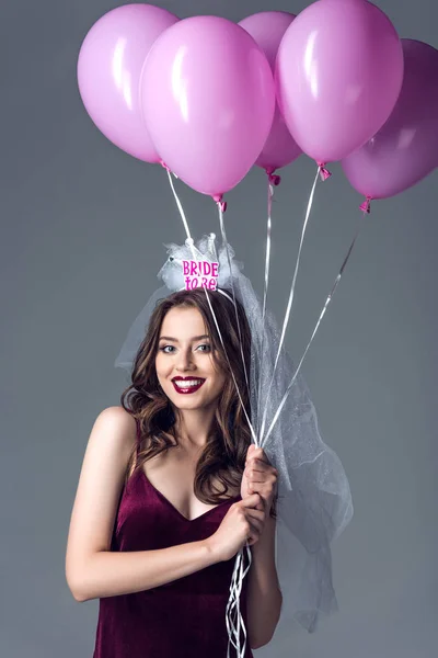 Усміхнена майбутня наречена в завісі для дівич-вечора тримає купу рожевих повітряних кульок ізольовані на сірому — стокове фото