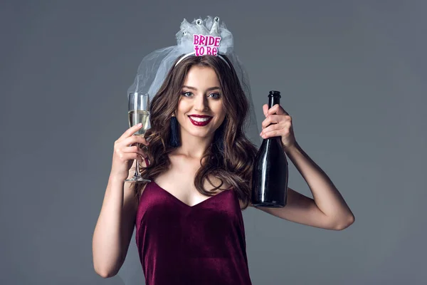 Beautiful future bride in veil for bachelorette party with champagne bottle and glass looking at camera isolated on grey — Stock Photo