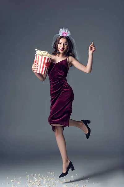 Beautiful future bride in veil for bachelorette party jumping with bucket of popcorn on grey — Stock Photo