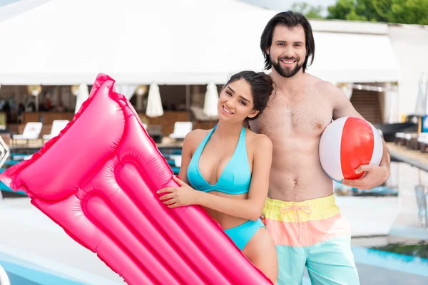 Couple swimsuits posing with inflatable mattress and ball near swimming pool — Stock Photo