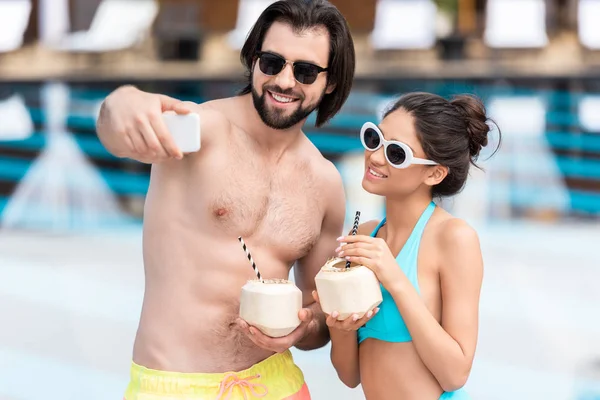 Young couple in sunglasses with coconut cocktails taking selfie on smartphone — Stock Photo