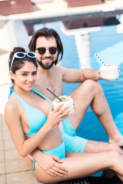 Jovem casal em óculos de sol com coquetéis de coco relaxante na piscina — Fotografia de Stock