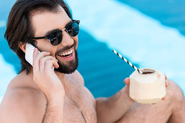 Homme souriant avec cocktail de noix de coco parler sur smartphone au bord de la piscine — Photo de stock