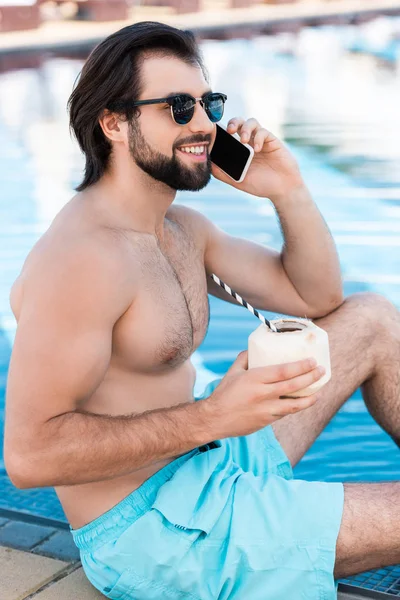 Bel homme avec cocktail de noix de coco parlant sur smartphone à la piscine — Photo de stock