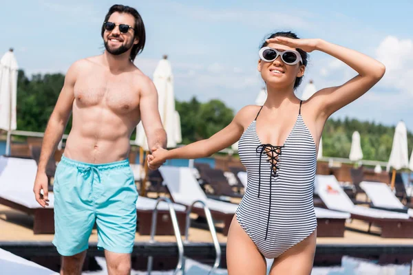 Novio y novia en gafas de sol cogidas de la mano cerca de la piscina - foto de stock