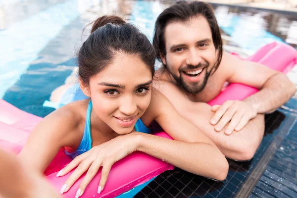 Красивая улыбающаяся пара плавает на розовом надувном матрасе в бассейне и делает селфи — стоковое фото