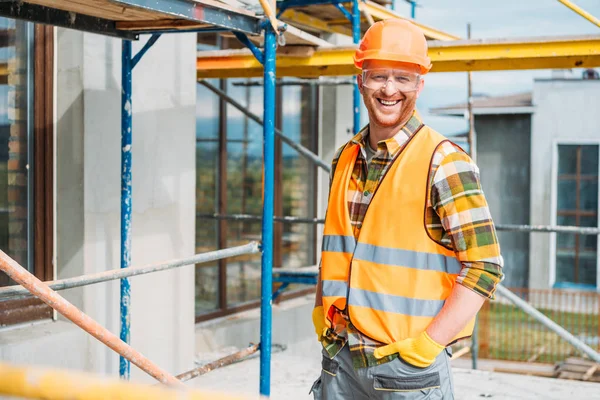 Beau constructeur souriant en gilet réfléchissant et chapeau dur regardant la caméra sur le chantier de construction — Photo de stock