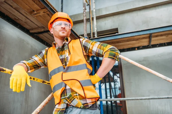 Beau constructeur appuyé sur échafaudage sur le chantier de construction et regardant loin — Photo de stock