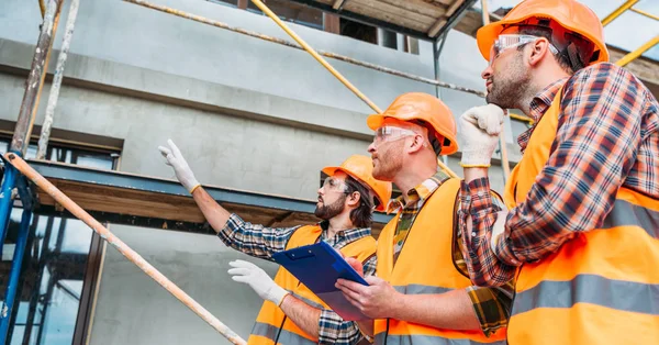 Wide shot of group of builders in hard hats and reflective vests pointing at building house — Stock Photo