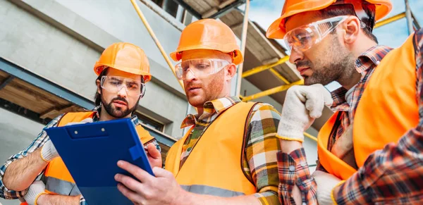 Wide shot of group of builders in hard hats and reflective vests looking at clipboard — Stock Photo
