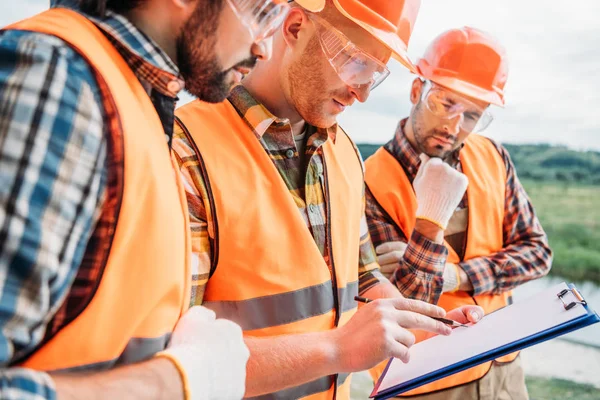 Group of builders in hard hats and reflective vests looking at clipboard — Stock Photo