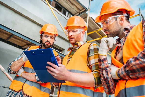 Bottom view of group of builders in hard hats and reflective vests looking at clipboard — Stock Photo