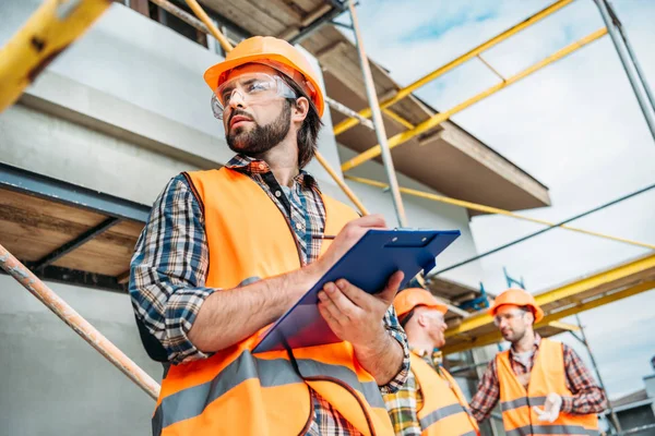 Blick von unten auf den gutaussehenden Bauunternehmer mit Klemmbrett, der wegschaut, während seine Kollegen verschwommen auf dem Hintergrund stehen — Stockfoto