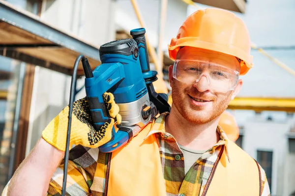 Apuesto constructor sonriente sosteniendo taladro de energía en el hombro y mirando a la cámara - foto de stock