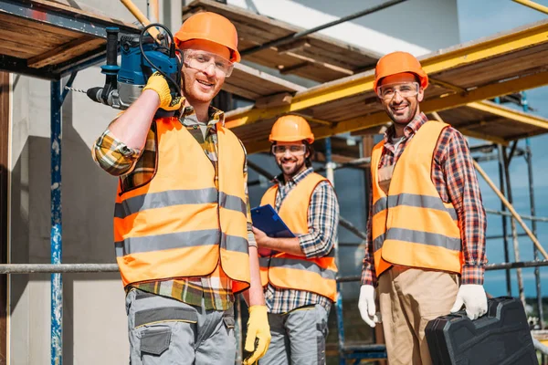 Grupo de construtores sorridentes com equipamentos de construção em pé no local de construção e olhando para a câmera — Fotografia de Stock