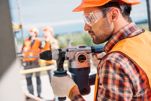 Vista laterale del costruttore in casco protettivo e occhiali da vista utilizzando trapano elettrico in cantiere — Foto stock