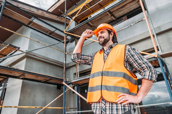 Construtor feliz em colete reflexivo e capacete em pé na frente da casa de construção e olhando para longe — Fotografia de Stock