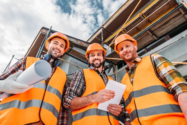 Bottom view of group of builders with blueprint and tablet looking at camera at construction site — Stock Photo