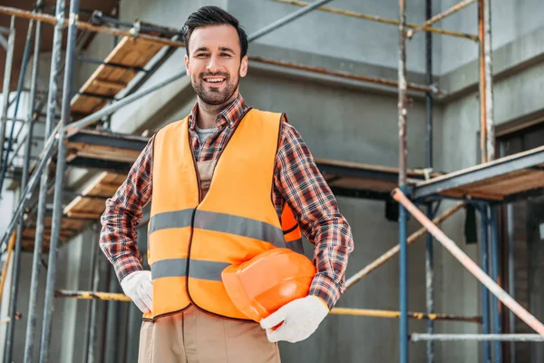 Happy builder in reflective vest holding helmet while standing in front of building house and looking at camera — Stock Photo
