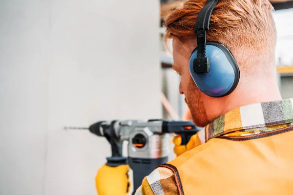 Confident builder in noise reducing headphones using power drill at construction site — Stock Photo