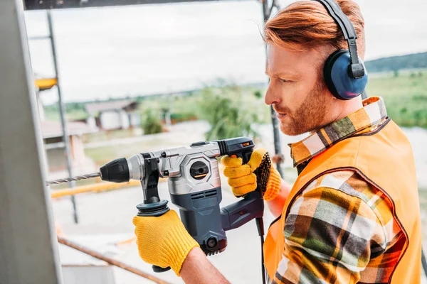 Side view of handsome builder in noise reducing headphones using power drill at construction site — Stock Photo