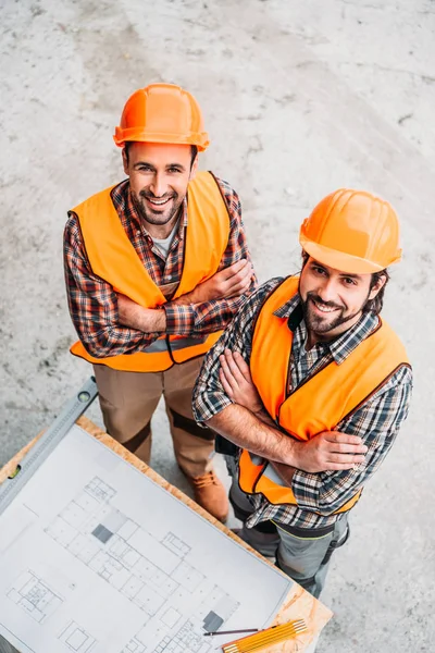 Vista ad alto angolo di costruttori felici con cianografia in piedi in cantiere e guardando la fotocamera — Foto stock