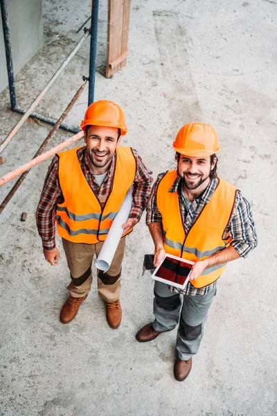 Vue grand angle des constructeurs avec plan et tablette debout sur le chantier de construction et regardant la caméra — Photo de stock