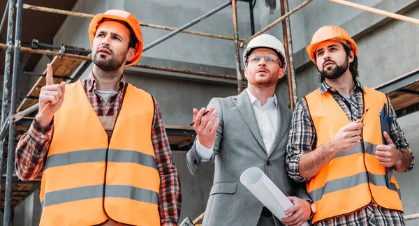 Tiro largo de construtores e arquiteto olhando para longe na frente do canteiro de obras — Fotografia de Stock