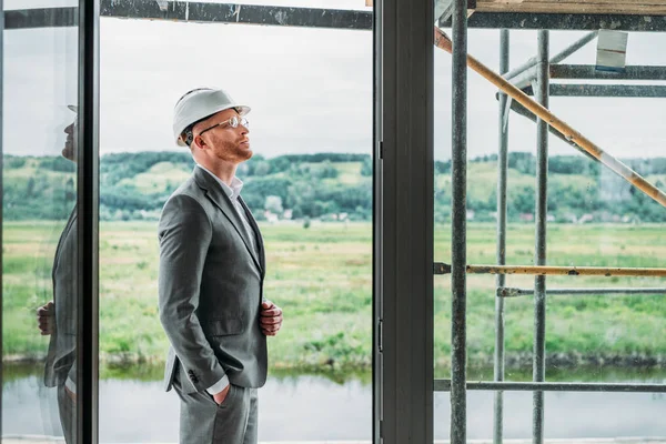 Side view of handsome architect in suit and hard hat standing on terrace at construction site — Stock Photo