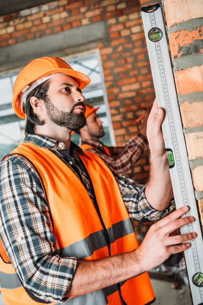 Vista laterale del bel costruttore utilizzando il livello della bolla in cantiere mentre il suo collega di lavoro su sfondo — Foto stock