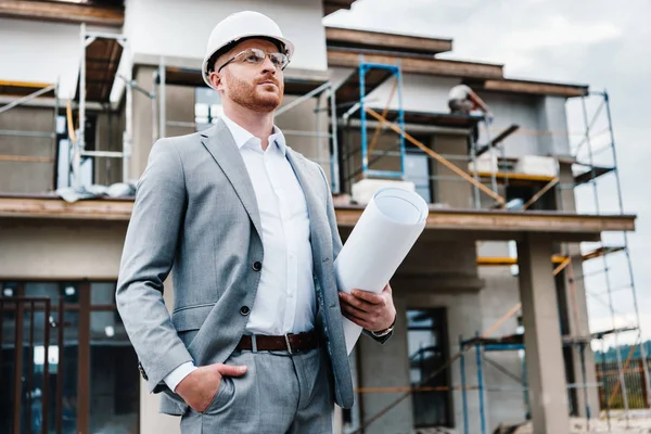Handsome architect in suit and hard hat holding blueprint standing in front of building house and looking away — Stock Photo