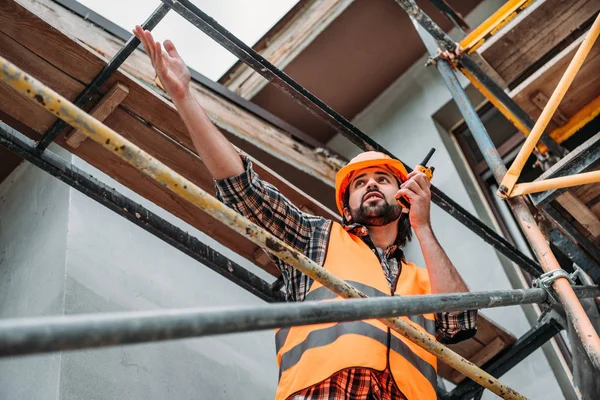 Vista inferior del constructor en casco protector y chaleco usando walkie talkie en el sitio de construcción - foto de stock