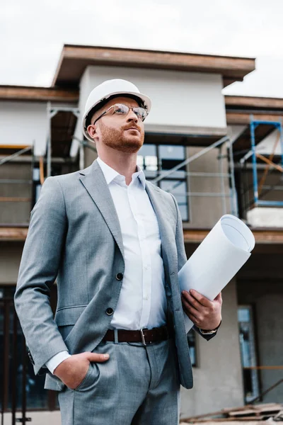 Handsome architect in suit and hard hat holding blueprint standing in front of building house — Stock Photo