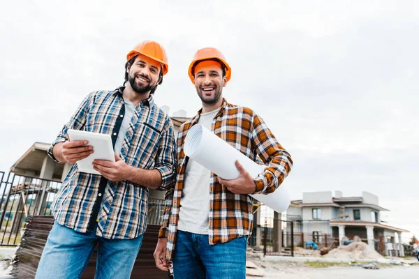 Architects with blueprint and tablet looking at camera at construction site — Stock Photo