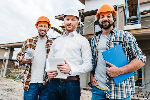 Gruppe lächelnder Architekten mit harten Hüten, die vor dem Haus in die Kamera schauen — Stockfoto