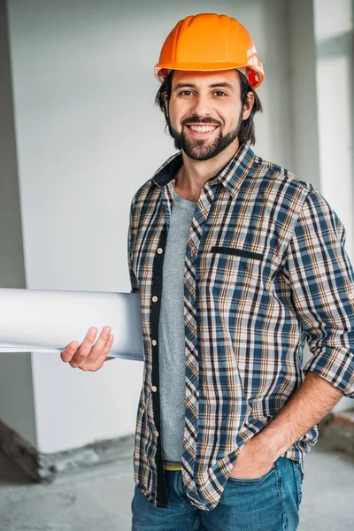 Erfolgreicher lächelnder Architekt in kariertem Hemd und harter Mütze steht mit Blaupause im Haus und blickt in die Kamera — Stockfoto