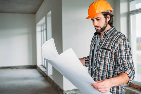 Apuesto arquitecto sonriente en camisa a cuadros y sombrero duro de pie dentro de la casa del edificio con plano - foto de stock