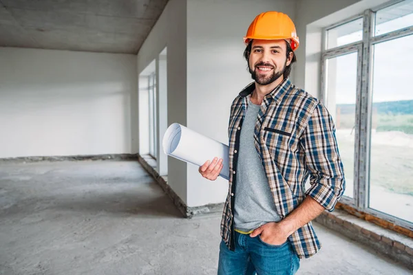 Bonito sorrindo arquiteto em camisa xadrez e chapéu duro de pé dentro da casa de construção com planta e olhando para a câmera — Fotografia de Stock