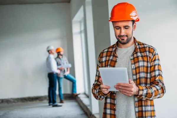 Bel architecte en chemise à carreaux et chapeau dur debout à l'intérieur de la maison de construction et en utilisant une tablette — Photo de stock