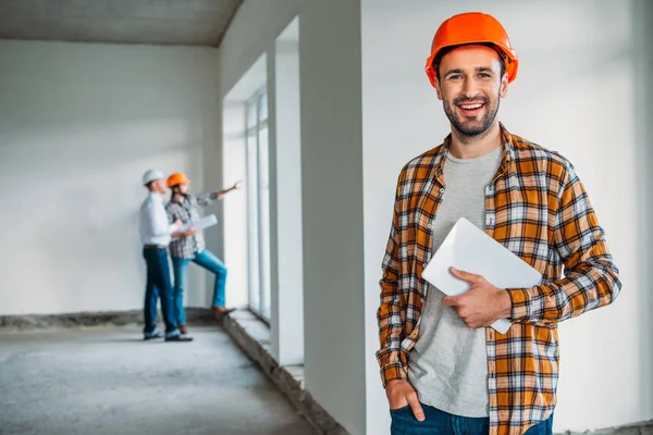 Arquiteto bonito em camisa xadrez e chapéu duro em pé dentro da casa de construção com tablet nas mãos — Fotografia de Stock