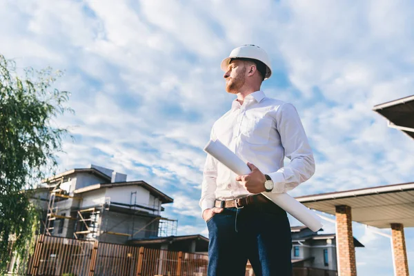 Bottom view of handsome architect in white shirt with blueprint standing at yard in front of building house and looking away — Stock Photo
