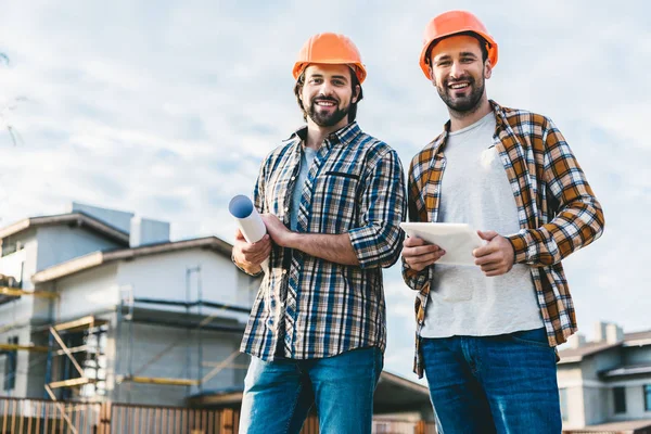 Architects with blueprint and tablet looking at camera at construction site — Stock Photo