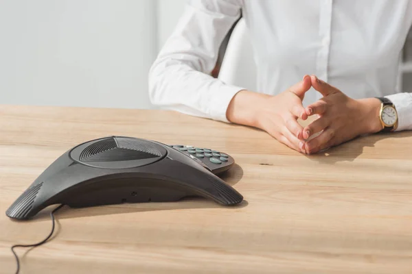 Cropped shot of businesswoman in white shirt sitting in front of conference phone on wooden table — Stock Photo