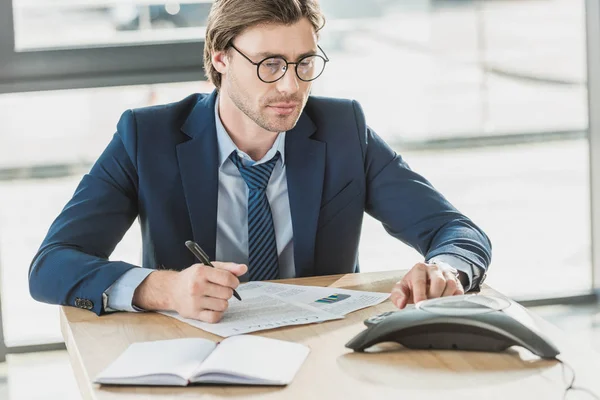 Bel giovane uomo d'affari con un sacco di scartoffie utilizzando il telefono conferenza in ufficio — Foto stock